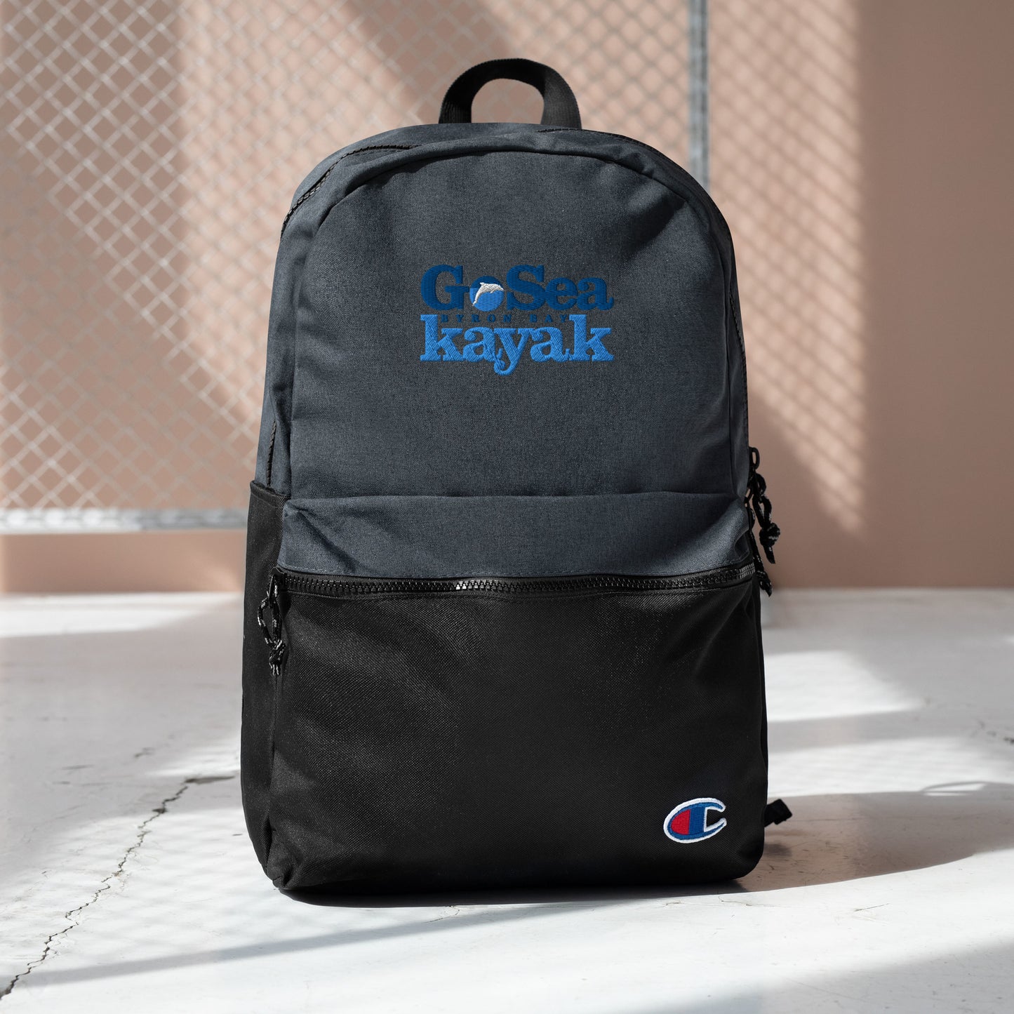 Champion Backpack - Black - Front view - Bag sitting on floor in front of fence - With Go Sea Kayak Byron Bay logo on front  - Genuine Byron Bay Merchandise | Produced by Go Sea Kayak Byron Bay 