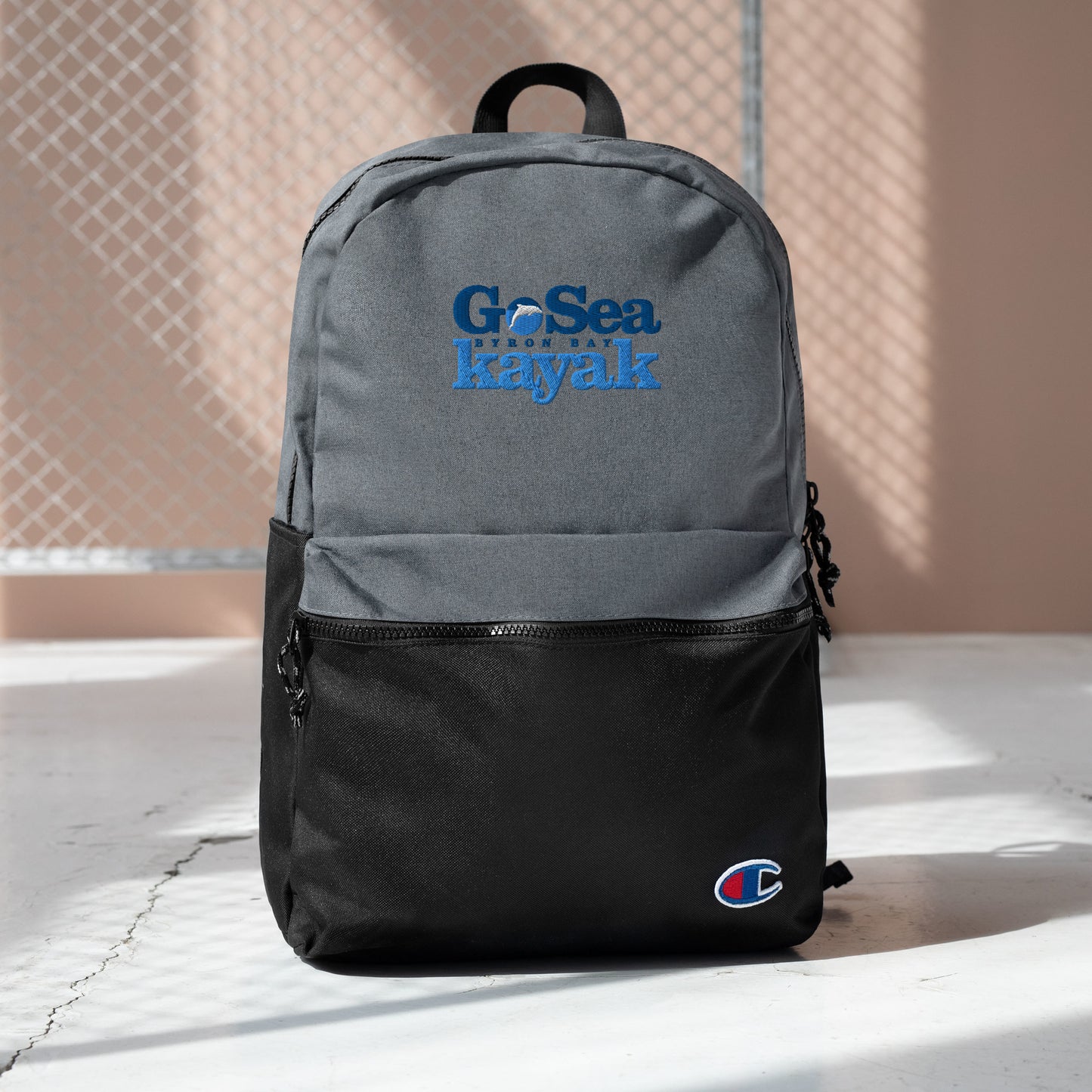  Champion Backpack - Grey and Black - Front view - Bag sitting on floor in front of fence - With Go Sea Kayak Byron Bay logo on front  - Genuine Byron Bay Merchandise | Produced by Go Sea Kayak Byron Bay 