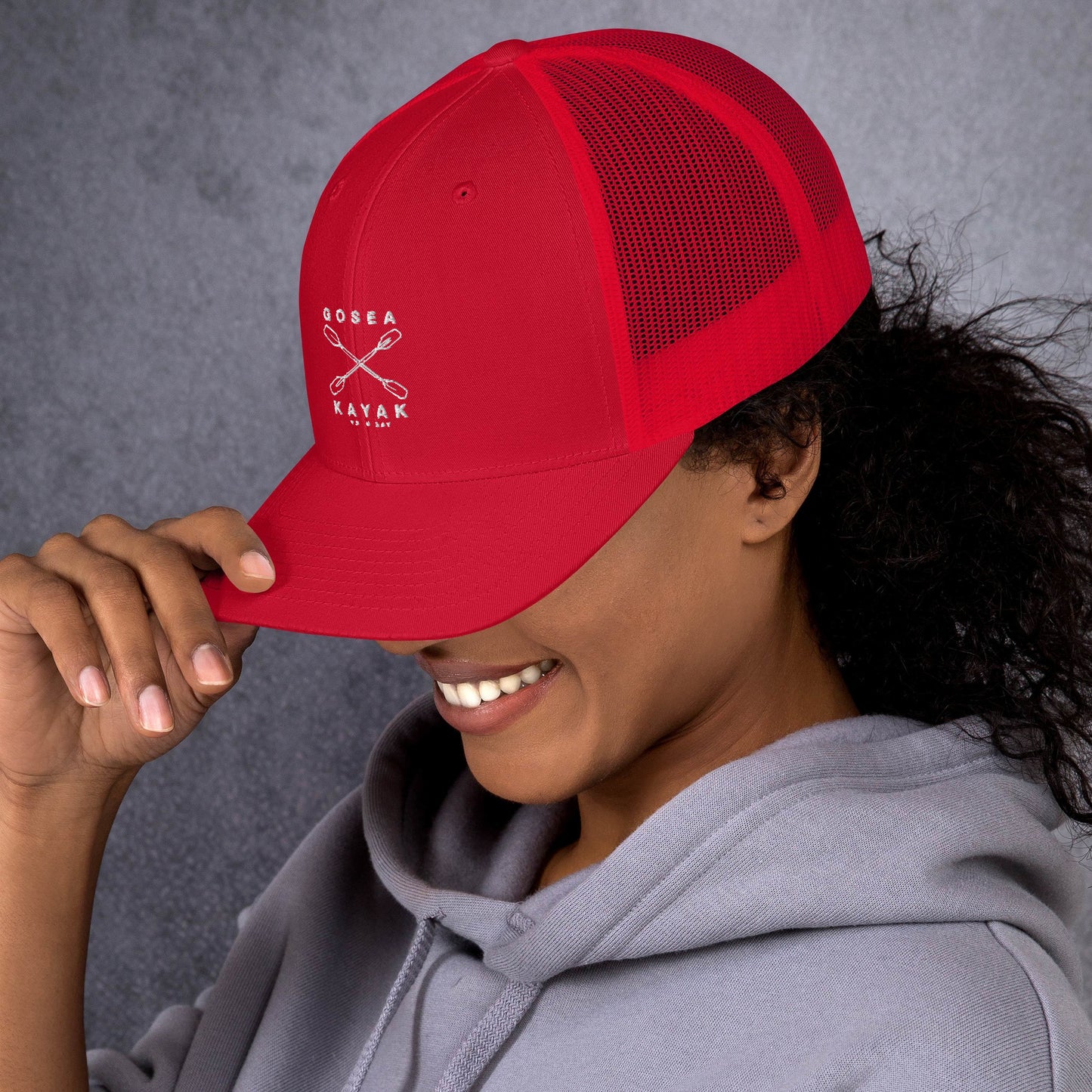  Trucker Cap - Red - side view - being warn by woman with her head down and hand holding the brim - Crossed Paddles Go Sea Kayak Crew logo in white on front - Genuine Byron Bay Merchandise | Produced by Go Sea Kayak Byron Bay 