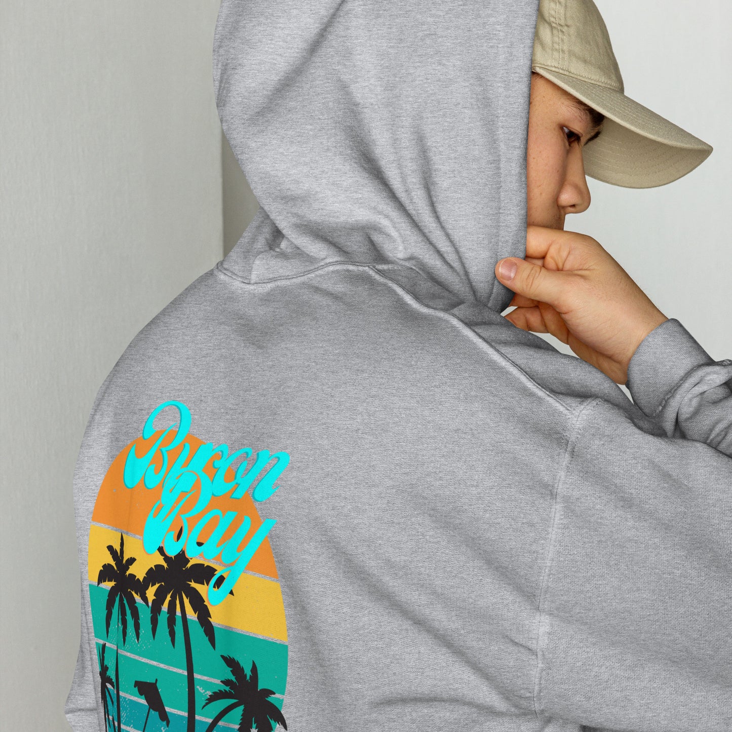  Unisex Hoodie - Sport Grey - Side back view being warn by man with the hood on looking back over his shoulder - Byron Bay design on back, plain front with pouch pocket - Genuine Byron Bay Merchandise | Produced by Go Sea Kayak Byron Bay 